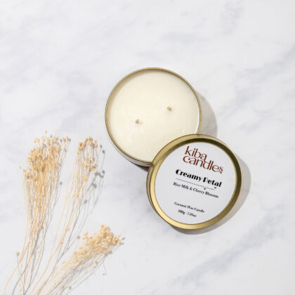 A creamy petal candle from Kiba, featuring a scent of rice milk and cherry blossom, presented in a round tin with a gold lid, placed on a marble surface alongside dried flowers.