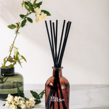 A dark amber diffuser bottle with black reeds, surrounded by green foliage and small white flowers, on a marble surface.