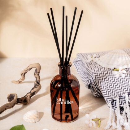 A decorative amber glass bottle with black reed sticks, labeled 'kiba candles', set against a light background with shells and a textured cloth.