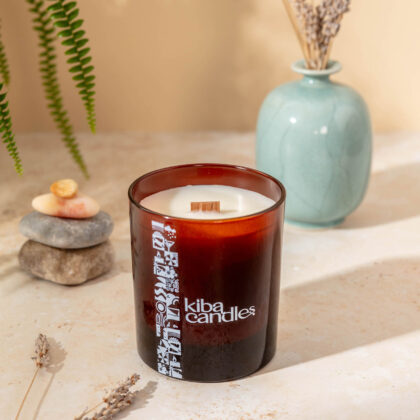 A Kiba scented candle in an amber glass container with a wooden wick, placed on a stone surface next to pebbles and a vase with dried flowers.