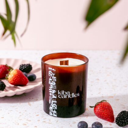 A Kiba scented candle in a brown glass jar with a wooden wick, surrounded by fresh strawberries and blackberries on a light speckled surface.