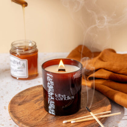 A lit Kiba candle on a wooden plate with smoke rising, accompanied by a jar of honey and wooden matches, set against a neutral background.