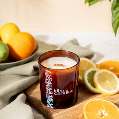 Coconut wax candle surrounded by fresh fruits, including oranges and lemons, placed on a wooden board with a green cloth backdrop.