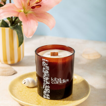 A Kiba scented candle displayed on a decorative plate with seashells, surrounded by a flower and a striped vase, against a soft blue background.