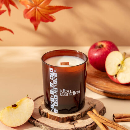 Coconut wax candle with a wooden wick, surrounded by fresh apple slices and cinnamon sticks, on a wooden surface with autumn leaves.