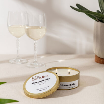 A coconut wax candle labeled 'Vineyard Mist' sits open on a neutral tablecloth, alongside two glasses of white wine and a small plant in the background.